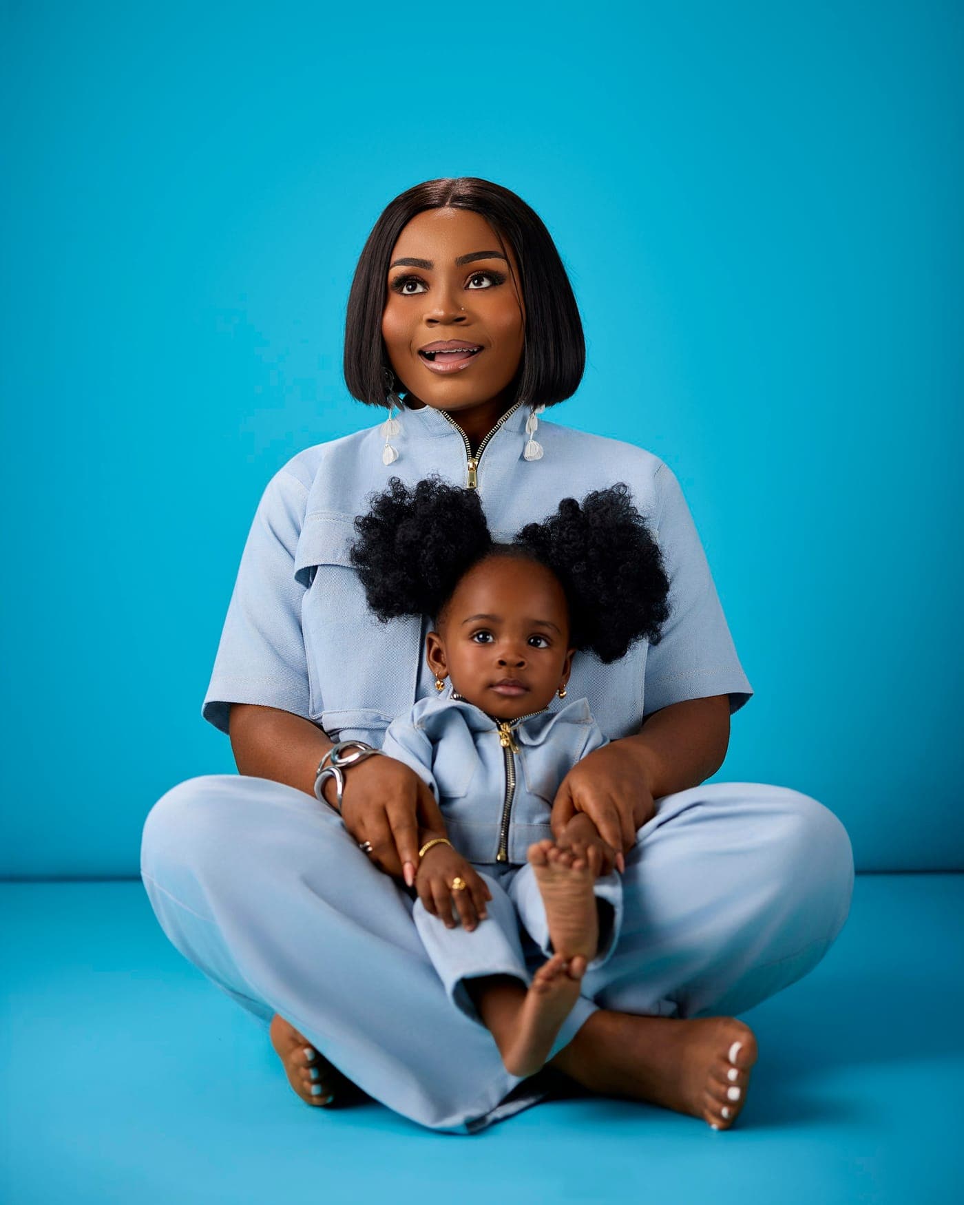 A smiling mother sitting cross-legged with her baby daughter on her lap against a sky-blue backdrop